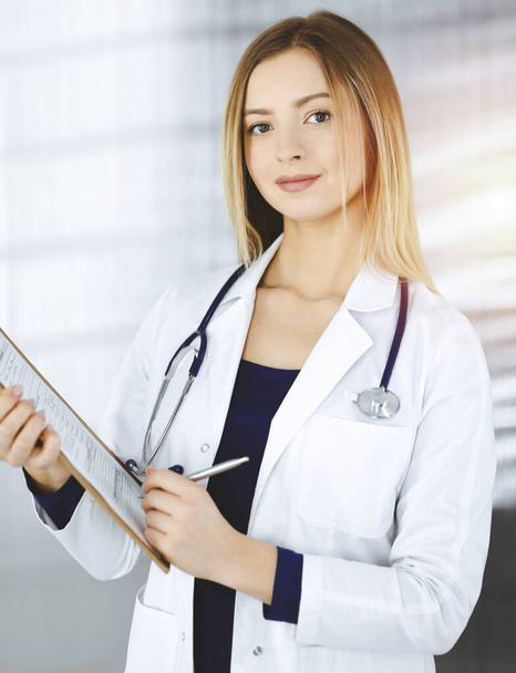 Young optimistic woman-doctor is holding a clipboard in her hands, while standing in a sunny clinic. Portrait of friendly female physician with a stethoscope. Perfect medical service in a hospital - 写真・画像