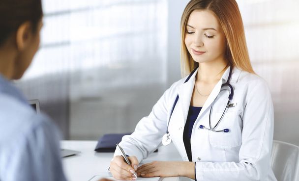 Unknown woman-doctor is writing some medical recommendations to her patient, while they are sitting together at the desk in the sunny cabinet in a clinic. Physician is using a clipboard, close-up - Foto, Imagen
