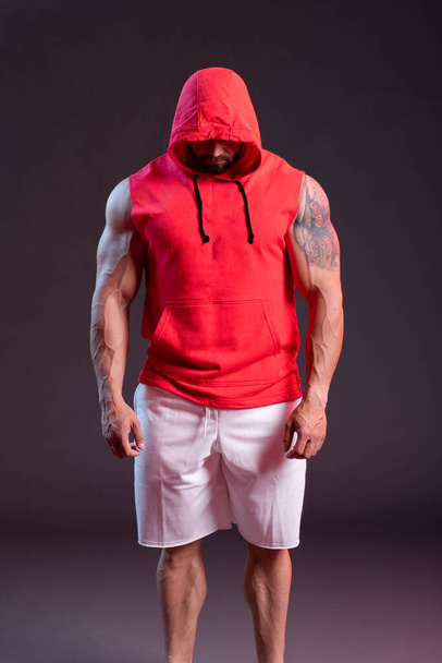 Powerful muscular man with a hood posing in the red sweatshirt. Studio shoot with red and blue smoke - Фото, изображение