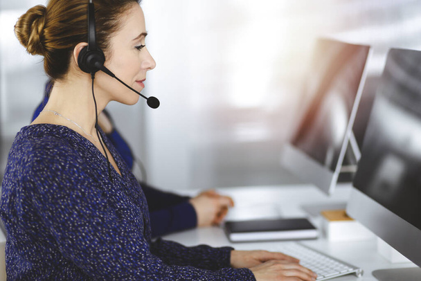 Two busineswomen have conversations with the clients by headsets, while sitting at the desk in a sunny modern office. Diverse people group in a call center - Foto, imagen
