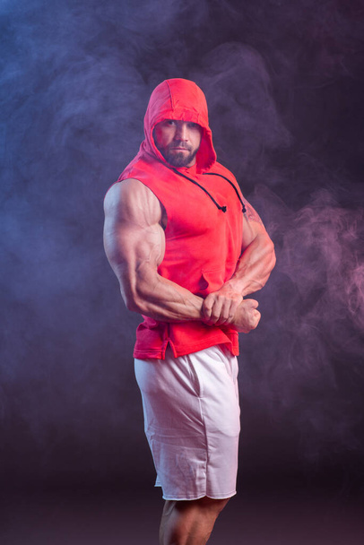 Powerful muscular man with a hood posing in the red sweatshirt. Studio shoot with red and blue smoke - Fotografie, Obrázek