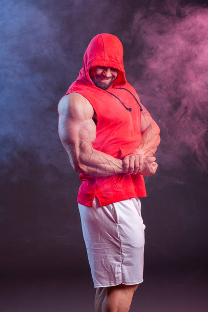 Powerful muscular man with a hood posing in the red sweatshirt. Studio shoot with red and blue smoke - Foto, Imagem