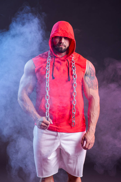 Powerful muscular man with a hood and chain posing in the red sweatshirt. Studio shoot with red and blue smoke - Photo, image