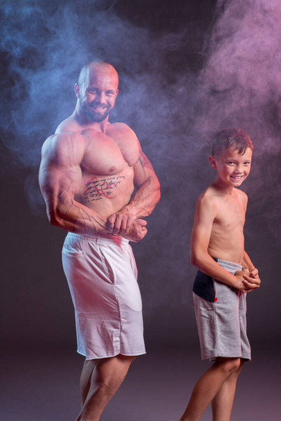 Powerful muscular man shirtless posing with his son in the studio with red and blue smoke - Foto, Bild