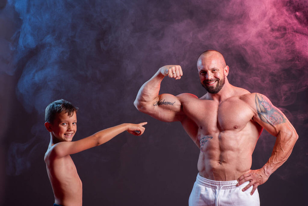 Powerful muscular man shirtless posing with his son in the studio with red and blue smoke. Little boy showing biceps contraction - Foto, Bild