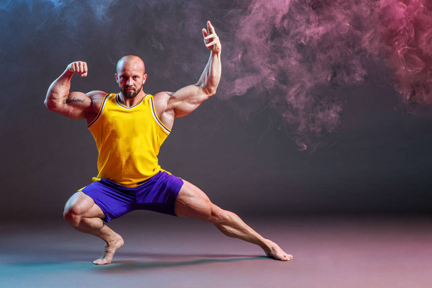 Mocný svalnatý muž pózující ve studiu s červeným a modrým kouřem, bicepsy a kontrakcí nohou - Fotografie, Obrázek
