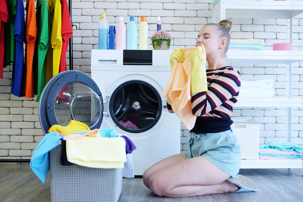 Lächelnde junge Kaukasierin Tragen Sie gelbe Gummihandschuhe für saubere Hausarbeit in der Waschküche. Sie hält ein Handtuch in der Hand und riecht duftend mit Kleidungsstücken im Korb. Konzept "Familie im Urlaub". - Foto, Bild