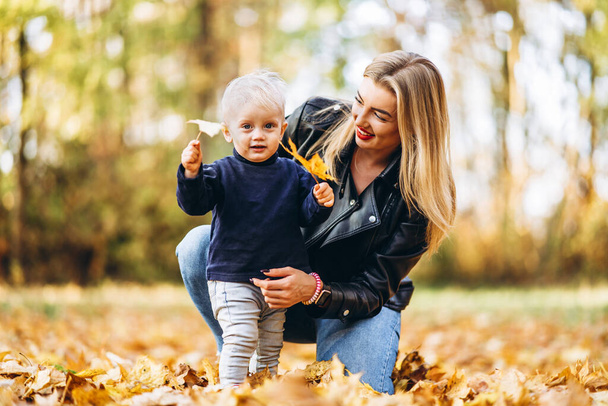 Happy mother with her little baby son have fun in the city park with colorful leaves around in autumn time. Happy family and mothers day concept - Fotografie, Obrázek