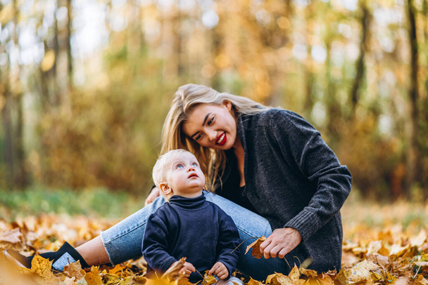 Happy mother with her little baby son have fun in the city park with colorful leaves around in autumn time. Happy family and mothers day concept - Photo, Image
