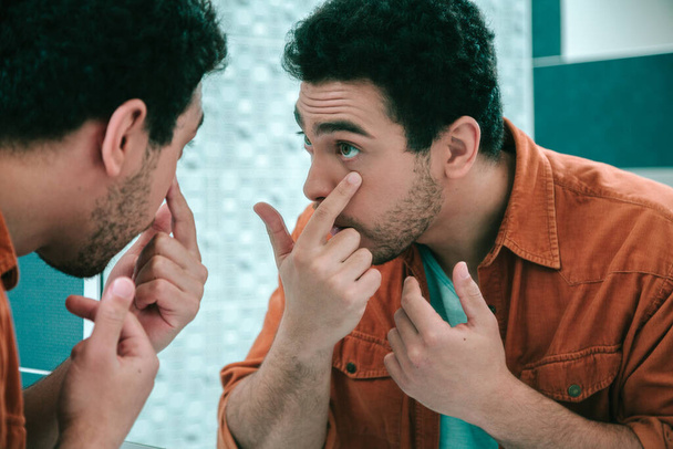 Careful young man standing in front of a mirror and pulling one lower eyelid further down with his finger - Photo, Image