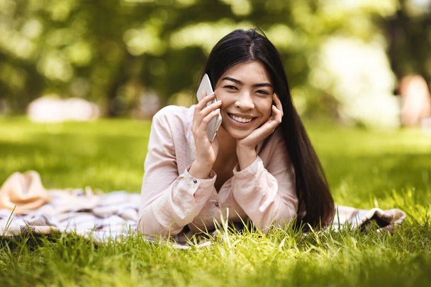 Ευχάριστο τηλεφώνημα. Ευτυχισμένη Κορεάτισσα που μιλάει στο κινητό ενώ χαλαρώνει στο πάρκο - Φωτογραφία, εικόνα