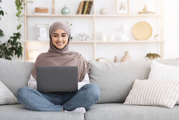 オンライン教育。自宅でソファの上でラップトップを使用してスカーフでアラビア語の女の子 - 写真・画像