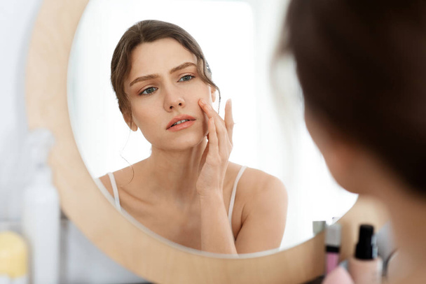 Расстроенная молодая женщина смотрит в зеркало, проверяет кожу на лице - Фото, изображение