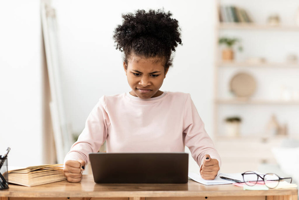 Θυμωμένο μαύρο κορίτσι κοιτάζοντας οθόνη φορητού υπολογιστή σφίγγοντας τις γροθιές στο εσωτερικό - Φωτογραφία, εικόνα