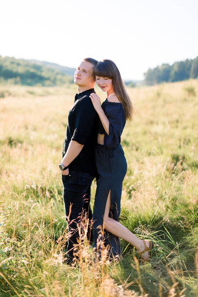 Young cute couple in love, wearing stylish black clothes, posing outdoor in summer meadow. Sensual romantic portrait of young stylish fashion couple in field, woman hugs his man from back - Foto, Imagem