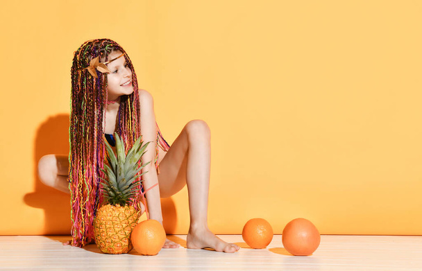 Teenage female with african pigtails, in swimwear. Squatting and smiling, posing with pineapple and oranges on yellow background - Photo, image