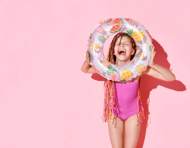 Cute girl z wielokolorowych afro kiści w jasnym różowym stroju kąpielowym śmiejąc się szczęśliwie trzymając gumowy pierścień w rękach. Trzy czwarte strzału na różowo - Zdjęcie, obraz