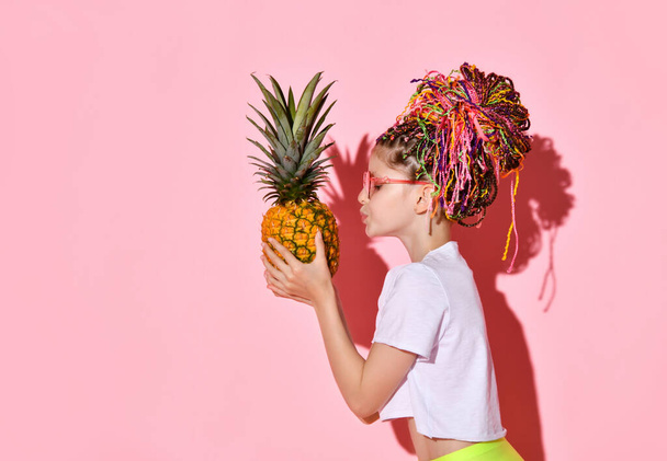 Side shot of cute girl with colorful braids in stylish red glasses looking on big pineapple in hands and going to kiss it, isolated on pink - Fotoğraf, Görsel