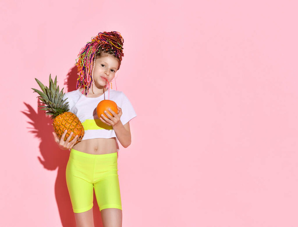 cute little girl with multi-colored pigtails on her head smiles happily, holding a pineapple and an orange in her hands. Cropped portrait isolated on pink, copy space. Childhood, emotions, summer. - Photo, image