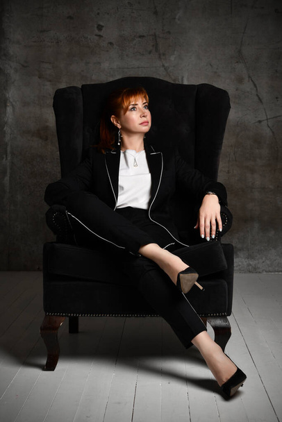 Thoughtful pretty woman in black trouser suit and high heels sits cross-legged in armchair at dark room pensively looking aside - Фото, зображення