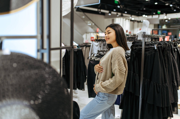 Młoda kobieta przymierza sweter w sklepie odzieżowym. Kobieta zakupy w butiku mody, zakupoholiczka, shopper patrząc na odzież - Zdjęcie, obraz
