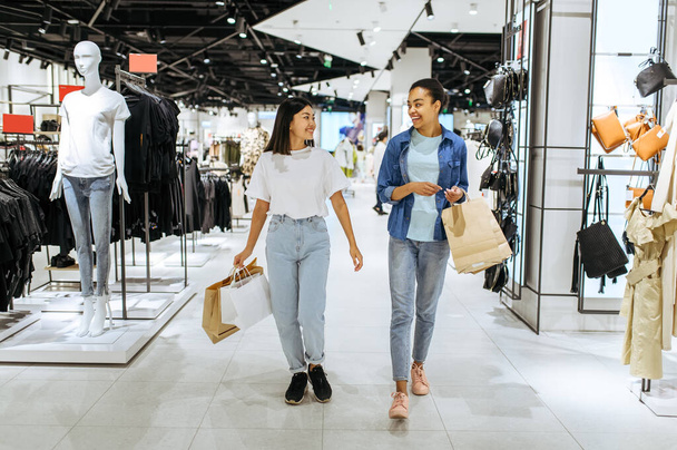 Smiling girlfriends with cardboard bags walks in clothing store. Women shopping in fashion boutique, shopaholics, shoppers with purchase - 写真・画像