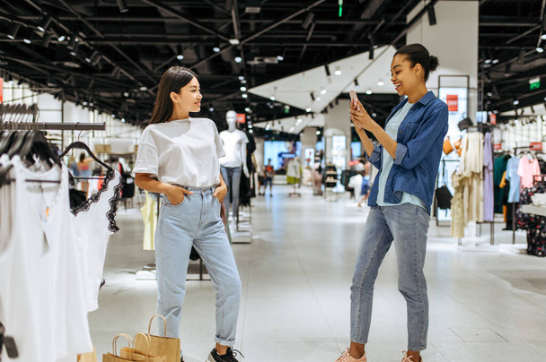 Lächelnde Freundinnen mit Pappbeuteln gehen in ein Bekleidungsgeschäft. Frauen shoppen in Modeboutique, Shopper mit Einkauf - Foto, Bild