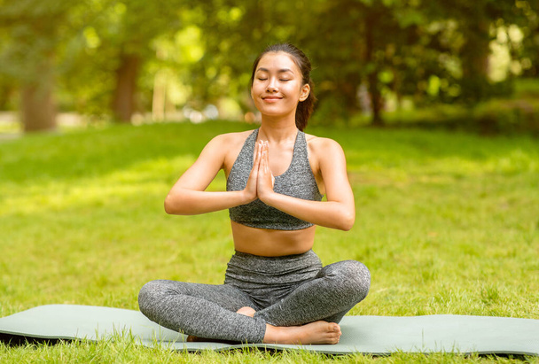 Asian girl sitting in lotus pose and doing yoga meditation or breathing exercises at park - Photo, Image