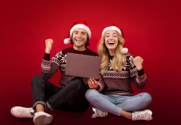 Full length portrait of millennial couple in Christmas clothes gesturing YES near laptop computer on red background - Photo, Image