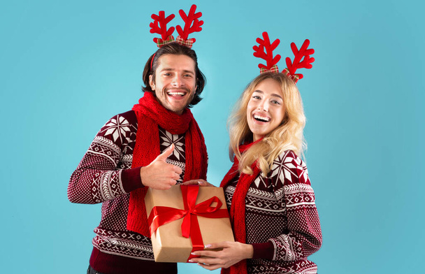 Romantisch jong paar in kerst outfits houden geschenkdoos en tonen duim omhoog gebaar op blauwe achtergrond - Foto, afbeelding