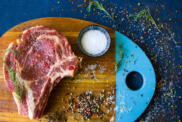 Rohes Rindfleisch auf dem Tisch, Gewürze und Rosmarin, Zutaten für Rib Eye Steak. Pfeffer und grobes Salz. Natürliches Licht. - Foto, Bild