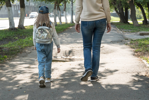 La madre lleva a la hija del niño a la escuela. Una chica con una mochila en la espalda está caminando con su madre en la calle. - Foto, imagen