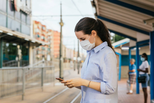 Archivbild einer jungen Frau mit Gesichtsmaske, die am Telefon plaudert, während sie an einer Straßenbahnhaltestelle wartet - Foto, Bild