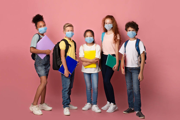 Edukacja podczas epidemii wirusa. Różnorodne dzieci z torbami szkolnymi i notatnikami w maskach na różowym tle - Zdjęcie, obraz