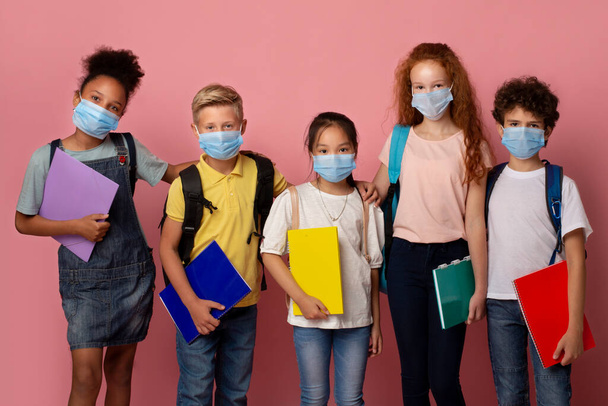 Vírus járvány és iskolázás. Többnemzetiségű gyerekek orvosi maszkok kezében notebook felett rózsaszín háttér - Fotó, kép