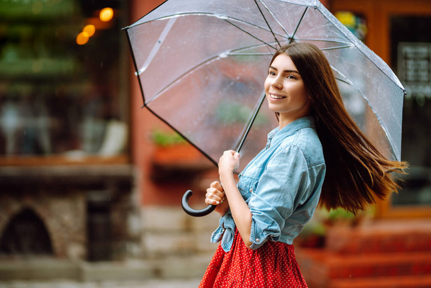 Vidám nő egy átlátszó esernyő alatt, aki élvezi az esőt. Egy fiatal nő rejtőzik az eső elől. Őszi koncepció. - Fotó, kép