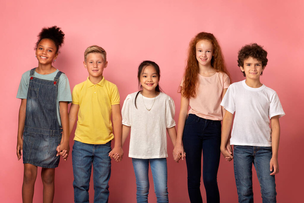 Glückliche multinationale Schulkinder, die sich vor rosa Hintergrund die Hände reichen - Foto, Bild