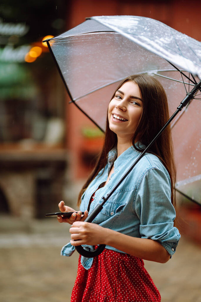Portrait of a smiling woman using phone under umbrella. Young woman with a transparent umbrella posing at city street. Autumn concept. - Фото, изображение