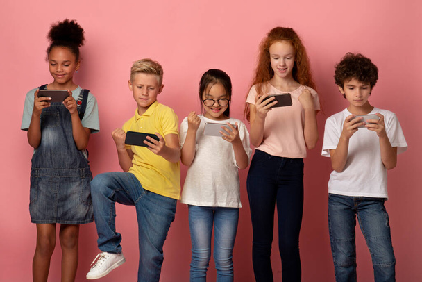 Cheerful schoolchildren playing mobile games on their phones over pink background - Foto, imagen