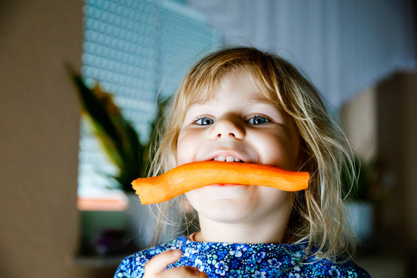 Söpö pikkulapsi tyttö pitelee ja puree tuoretta porkkanaa. Kauniilla lapsella on terveellinen välipala. Hymyilevä onnellinen lapsi syö bio orgaanisia vihanneksia - Valokuva, kuva