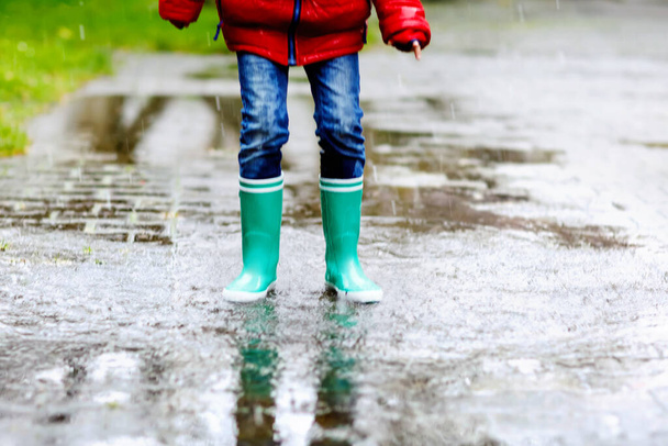 Sarı çizme giyen, yağmurda, yağmurda ve soğuk havada yürüyüş yapan çocuğun yakın çekimi. Renkli moda kıyafetleri içinde bir su birikintisine atlayan bir çocuk. Dışarıda eğleniyoruz. - Fotoğraf, Görsel