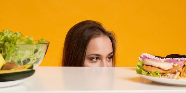 Жінка заглядає зі столу, вибираючи між здоровою та нездоровою їжею
 - Фото, зображення