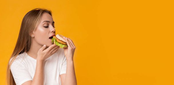 Нездорове харчування. Молода жінка їсть фастфуд, насолоджуючись смачним бургером
 - Фото, зображення