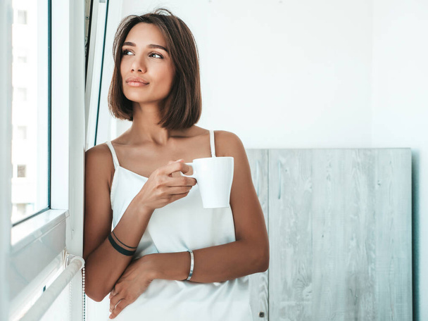 Portrait of beautiful woman dressed in white pajamas. Carefree female holds cup and drinking coffee. Sexy model enjoying her morning breakfast at balcony. She dreams about something - Fotó, kép