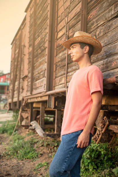 Joven con sombrero de vaquero mirando el atardecer frente a un vagón de tren de madera vintage. Fotografía del concepto de rancho - Foto, imagen