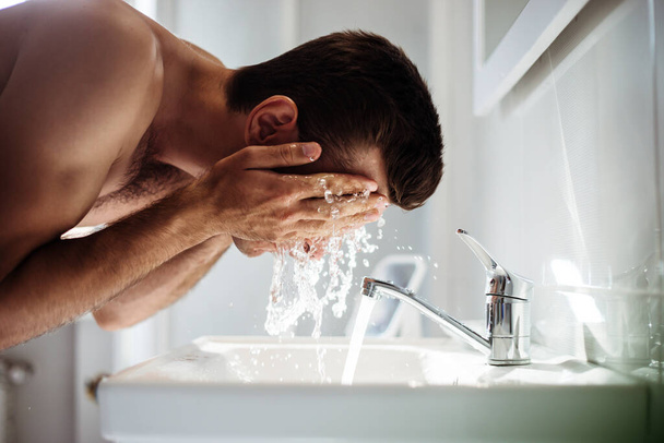 Handsome young naked man is washing his face in the bathroom at home. - Foto, Bild