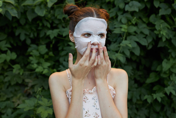 Nette Frau weiße Maske Hält seine Hände nah an sein Gesicht Hautpflege Büsche im Hintergrund - Foto, Bild