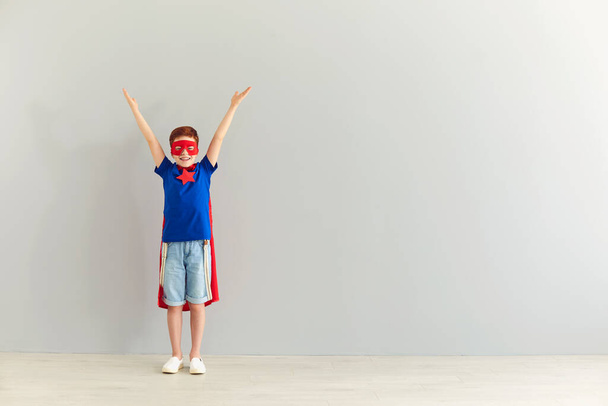 Smiling boy in costume of superhero standing with hands raised over grey background - Photo, image