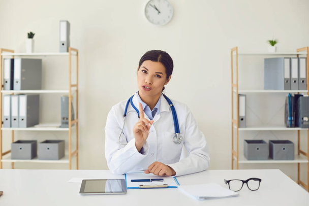 Doctor online. Hispanic female doctor looking at the camera says diagnose while sitting at a table in a clinic office - 写真・画像