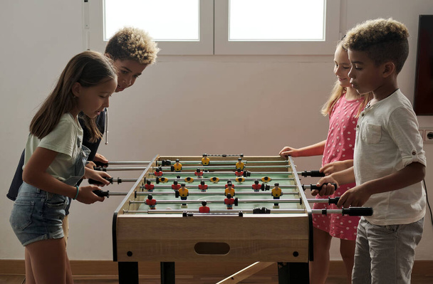 Group of multi cultural cute children stand indoor spend playtime with friends playing foosball together. Tabletop version of soccer lovers. Having fun and recreation of alpha generation kids concept - Fotoğraf, Görsel